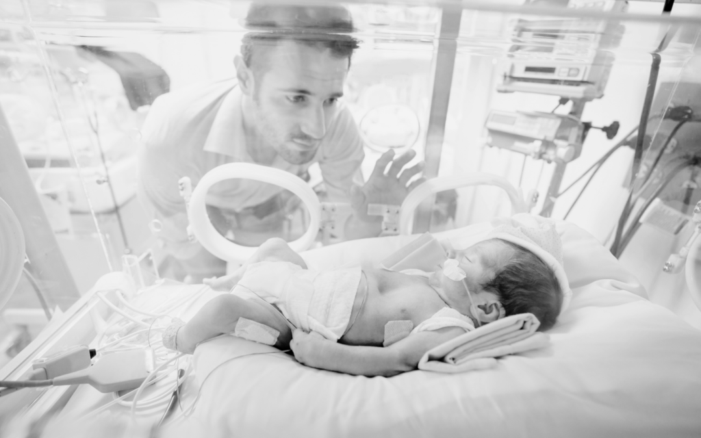 Baby lies in hospital incubator as father looks on with concern.